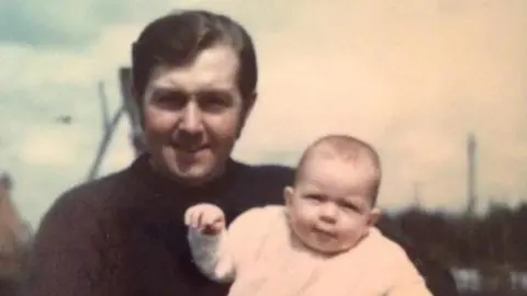 Melanie Stinton Stinton as a baby with her dad. Stinton is in a pale coloured baby grow, whereas her dad has short brown hair and a dark coloured top.