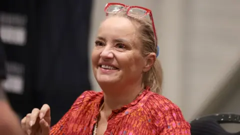 Lucy Davis, a fair-haired woman in a red top and with red-framed glasses worn on top of her head, is seen sitting down and smiling at the For The Love of Fantasy & Sci-Fi convention at Excel London on Saturday 22 August 2026