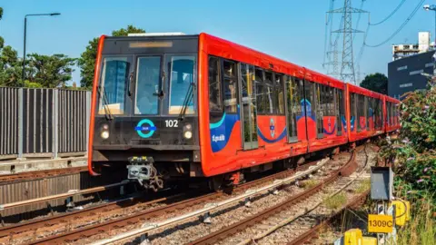 LightRocket via Getty Images A red DLR train moving on the tracks, viewed from the side, on a sunny day.