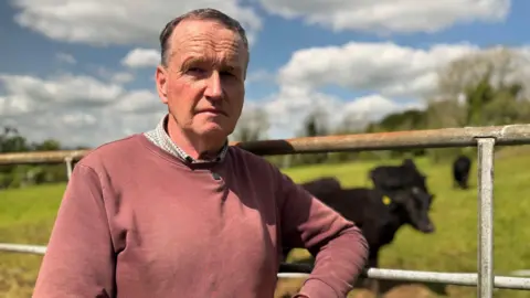 A man with short, greying hair looking to the camera. He is leaning against a metal farm gate on a sunny day. There are a number of black cows in the field behind him. He is wearing a pink jumper over a checked shirt.