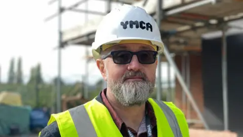 A man with a beard looking directly into the camera. He has a grey beard and dark glasses and is wearing a high vis jacket and hard hat. He is standing in front of a construction site covered in scaffolding.