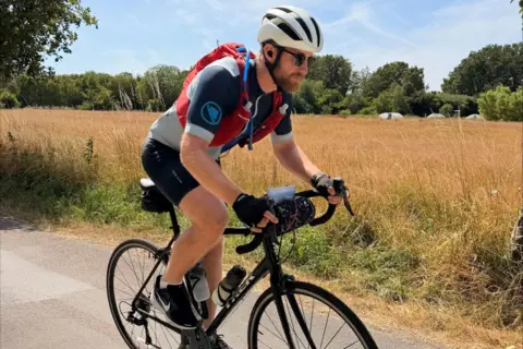 Matthew Hollingham A bearded man in cycling gear rides a bicycle on a road alongside fields and trees.