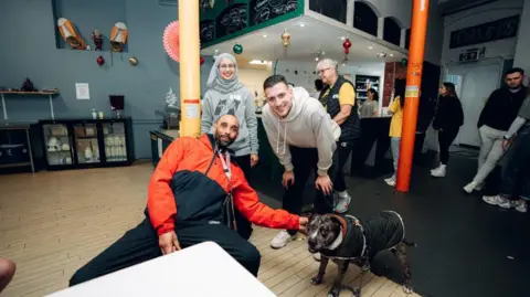 Diogo Dalot crouches over next to where a man is sitting on a chair holding the collar of a dog. A woman stands on the man's left. All three are smiling at the camera. 