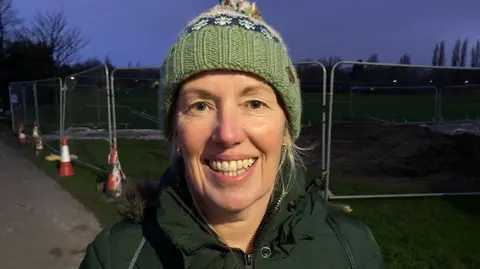 Close-up of Julie Norman who is smiling directly at the camera. She is wearing a green coat and woolly hat and is standing in front of metal barriers in a park. It is early morning and the sky is a dark purple.