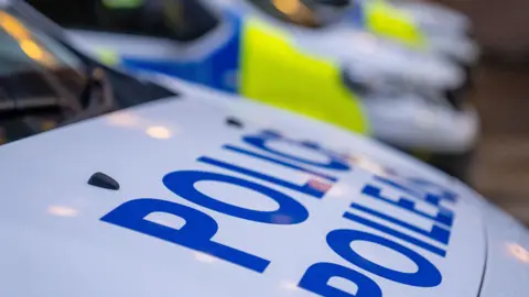 'Police' written in blue on white bonnet, with blue and yellow checks on vehicles in background.