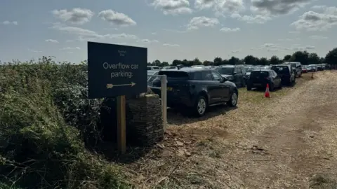 Lots of cars parked in a field. It's an over flow car park, according to the sign.