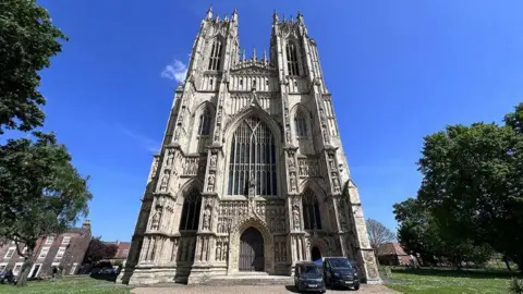 David Reeves/BBC A external shot of Beverley Minster. Two black vehicles are parked in front of the building facing the camera. Large green trees and grass surrounds the building. The sky is blue.