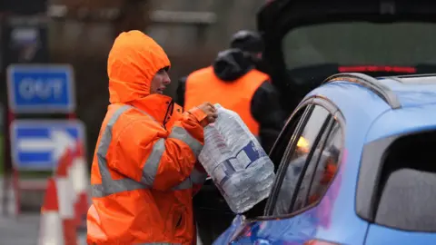 PA Media A woman in an orange jacket hands bottles of water through a car window. 
