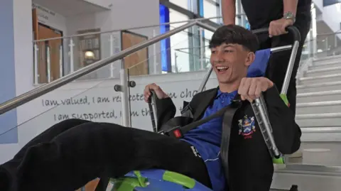 Lucas with brown hair wearing a school uniform smiles as he is lowered down the stairs of his school in an evacuation chair.