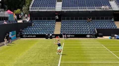 A female tennis player stands at the baseline on a grass tennis court, as she throws up the ball to serve. In the background there are banks of seats for spectators and there are also match officials standing to the left of the shot.