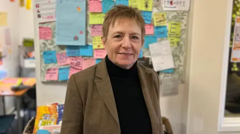 Victoria has short cropped auburn hair. She is wearing a brown blazer and black jumper. She is looking straight at the camera. In the background you can see multicoloured motivational post-it notes that young people have stuck up. 
