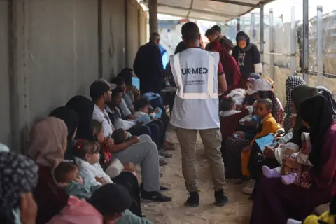 UK-Med Adults and children sitting in a corridor, with a man bearing a UK-Med vest in the middle.