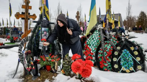 Reuters A woman puts a flower on a grave as she visits the Alley of Heroes at a local cemetery to pay tribute to victims of the Russian attack on Ukraine, on the day marking the fourth anniversary of the full-scale Russian invasion, in the town of Bucha, Kyiv region, Ukraine.