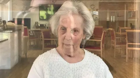 Jenny Harris Vivian Harris stares forward while sat in a chair inside a care home. She is wearing a white blouse and has grey hair. The reflection from a window can be seen in the image.