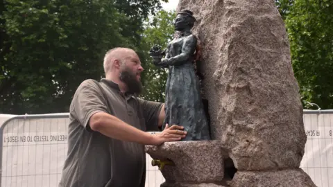 A man in a brown T-shirt installs a bronze statue on a large rock-style water fountain. There are tall trees in the background.