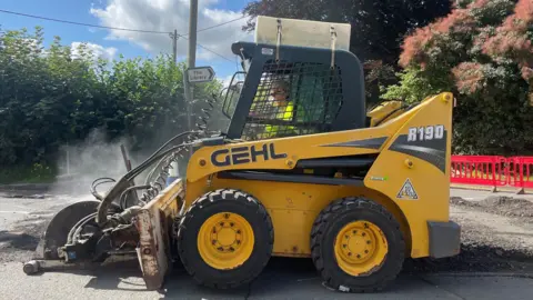 Gloucestershire County Council Small yellow vehicle with tractor tyres, a front attachment for folling potholes, and a caged cab for the driver