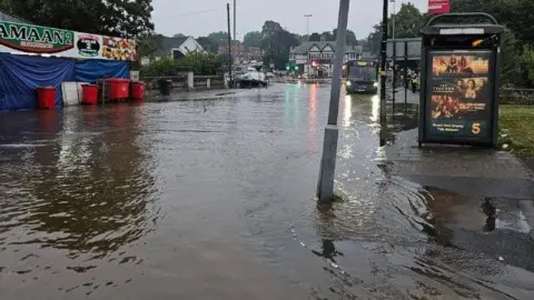 West Midlands Fire Service A flooded road with a bus stop and wheelie bins visible, there is a bus as well as various buildings in the background