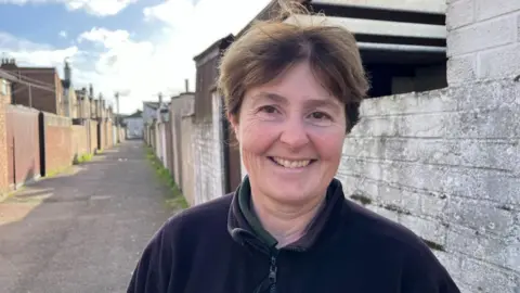 Andrew Turner/BBC Kerry Hemsworth, standing in an alley clear of rubbish and bins. She has auburn hair and wears a black fleece jacket.