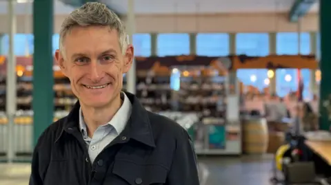 Ian Thorpe is standing in a shop with the background blurred. He has grey hair and is wearing a shirt and jacket and is smiling at the camera.