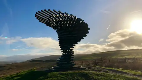 A sculpture made of pipes stacked on top of each other stands in the middle of green fields and fences with a sunset behind