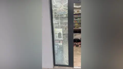Chaudhary Hamza Riaz The broken window of the shop. The window is in a grey frame and the glass is shattered.