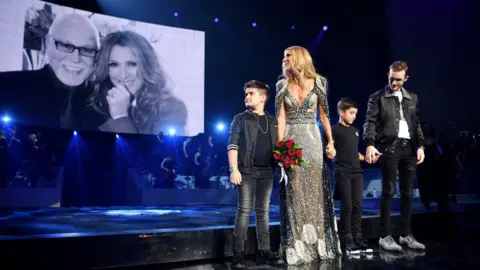 Getty Images Celine Dion holding hands with her children on stage, with a photo of her and late husband René Angélil projected on the screen