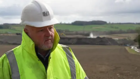 A man in a yellow hi-vis jacket and white hard hat stands on the quarry site 