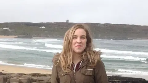 Pacemaker Natalie McNally. She has long blonde hair, wearing a long khaki coat and standing on a beach.