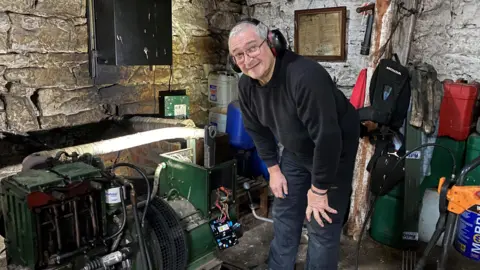 Claude Schiavetta is standing looking at a generator wearing headphones. He's a man in his sixties wearing a black jumper and jeans  