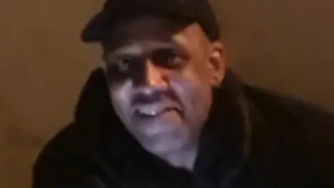 A man in low lighting smiles. He is wearing a black top and a black cap.