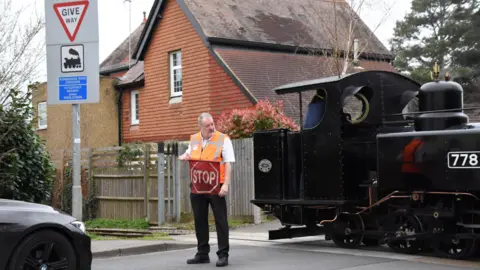 Leighton Buzzard Railway A man standing in a road, wearing an orange high-vis jacket, holding a red stop sign, by a train, with part of the front of a car to the left. A house is behind them. A sign is to the left and a fence behind the man. 