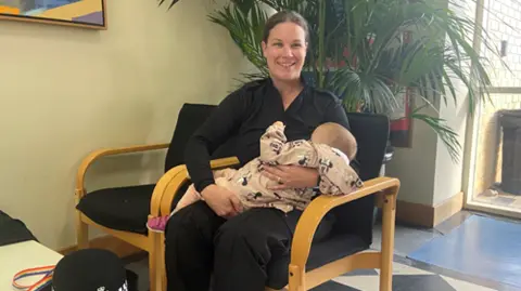 Sgt Kat Scrimshaw in a black shirt and black trousers. She is sitting in a chair. She is holding a small child in a pink onesie, and is breastfeeding. 