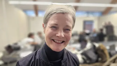 Margaret has light grey hair tied back behind her head. She is smiling at the camera and is wearing a navy polo neck and navy jacket.