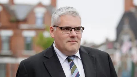 PA Media Gavin Robinson has grey hair and is wearing black-framed glasses. He is wearing a black suit jacket, white shirt and striped tie