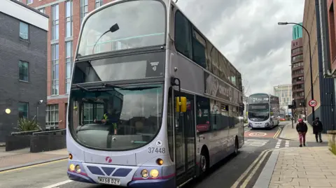 A First-branded purple double-decker bus travels along a road in Sheffield city centre, followed by another in the background.