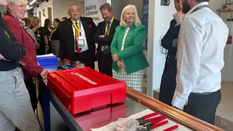 Emma Baugh/BBC A group of people inside the new facility. They are listening to a man talk. Next to him is a metal table with a red box on top as well as red pliers.