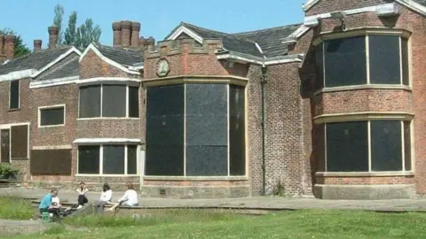 A photograph of Hopwood Hall in Middleton provided by Rochdale Council. It is an old brick building that has boarded up windows with a patch of untended grass at the front. A group of young people sit on the steps in front of it.