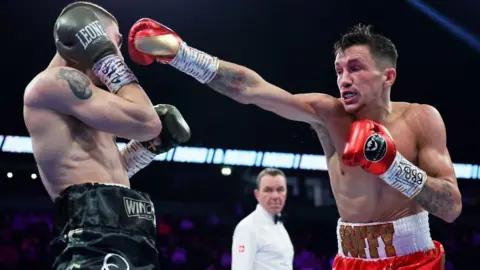 Getty Images Davies and another boxer are shirtless in a boxing ring. There is a referee blurred in the background. Davies has his arm outstretched in a punch, wearing red boxing gloves. The other boxer has his hands shielding his face and is wearing black shorts and boxing gloves.
