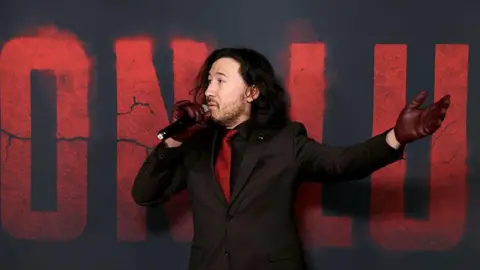 US YouTube star Mark Edward Fischbach stands on the red carpet for his debut film Iron Lung wearing a black suit with a red tie and with his medium length brown hair to his shoulders. 