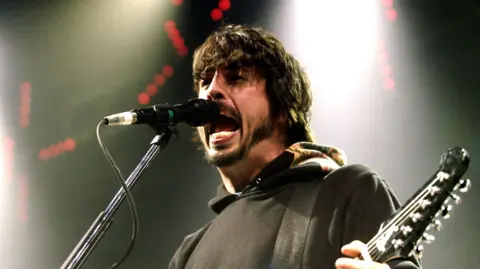 Dave Grohl sings into a microphone while playing a guitar. He has shoulder length thick black hair and a beard and is wearing a black hoody.