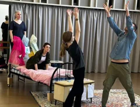 Sarah Lyle Cre8 actors rehearsing their play, Sleeping Beauty, ahead of the festival - woman on bed, woman stood beside her and male and female actors in front throwing books in the air