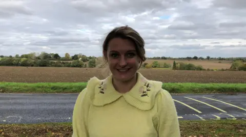 Vikki Irwin/BBC Ele Williamson smiles directly at the camera wearing a yellow jumper. She is photographed outside, with farmland behind her.