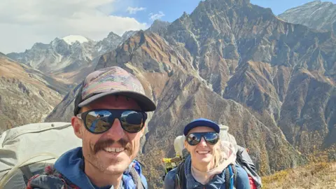 Rik de Louche Rik is taking a selfie of the couple and they're both smiling at the camera. They both have large backpacks on with blue fleeces and have sun glasses and caps on. Behind them is a mountain range.