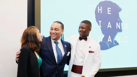 The Diana Award Alex Holmes, with his arms around a man and a woman, standing in front of a screen with The Diana Award printed on it. Alex is wearing a blue suit, with a white skirt and a blue tie, the woman has long hair, is wearing a green dress, with a jacket and the man on the right is wearing a white suit with a burgundy cumber band and bow tie. 