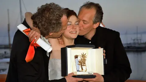 Getty Images Émilie Dequenne holding a Cannes Award with Luc and Jean-Pierre Dardenne on both sides kissing her on both cheeks in 1999