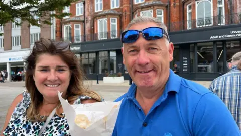 Andrew Turner/BBC Susie and Kenny Hearn, sitting in Great Yarmouth Market Place. She is wearing a peacock-tail patterned top, and has long brunette hair with her sunglasses on her head. Kenny is wearing a blue polo shirt, and has tinted sunglasses on his head. He is holding a cone of chips and gravy. Behind them is The Place, the new library and learning centre in Great Yarmouth Market Place.
