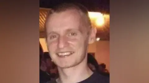 Devon and Cornwall Police Daniel Mizzi is smiling at the camera with people in the background and has light brown hair and is wearing a black T-shirt with red and yellow patterns on it