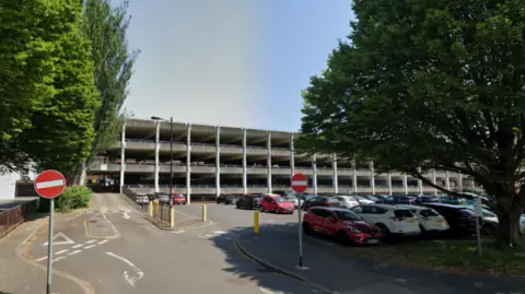 An image of a grey multi-storey car park with green trees either side
