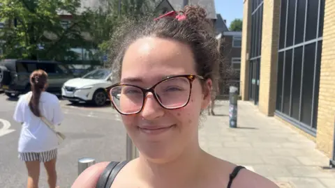 A woman with a black shirt, glasses and long, dark hair tied up. She is smiling at the camera and standing in a car park.