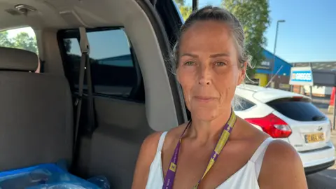 A woman in a white vest standing at the open boot of a van in a car park looking at the camera